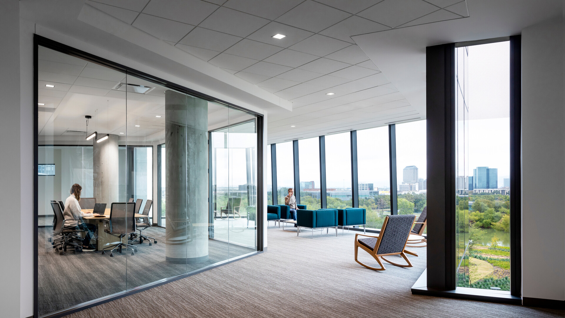 An open office space with carpeted floors, white ceiling tiles and large glass windows with a top-floor view